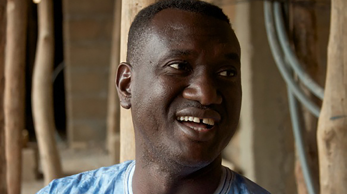 Bassekou Kouyate, wearing a blue top, smiles and in the background are roughly hewn wooden posts.